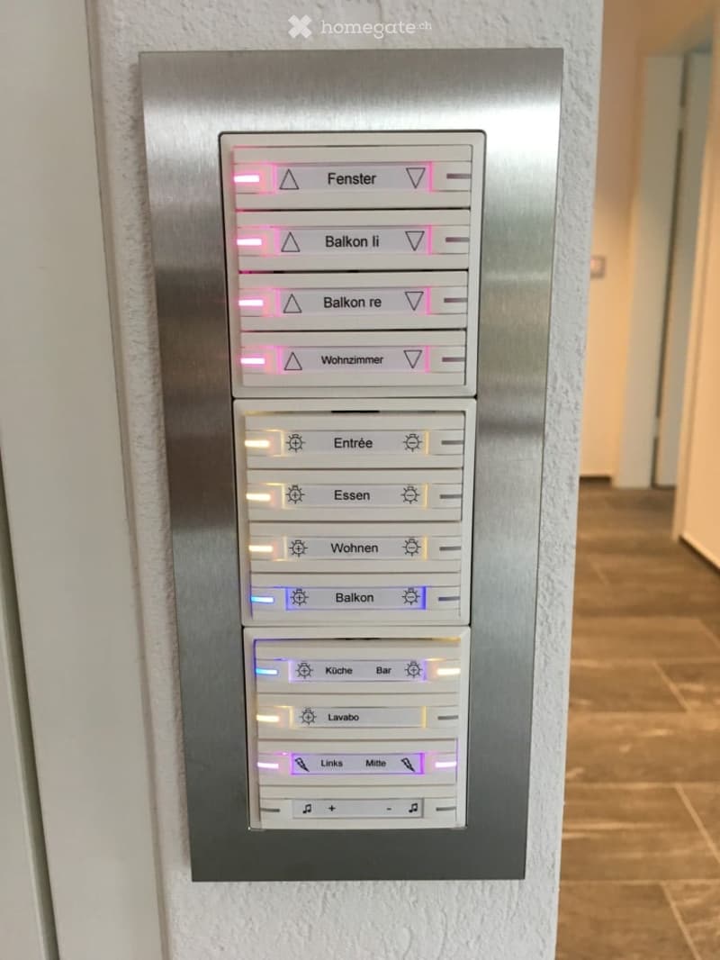 Modern control panel mounted on a wall in a hallway, featuring illuminated buttons for various functions such as windows, balconies, living room, entrance, dining, living, balcony, kitchen, bar, laundry, and room selection.