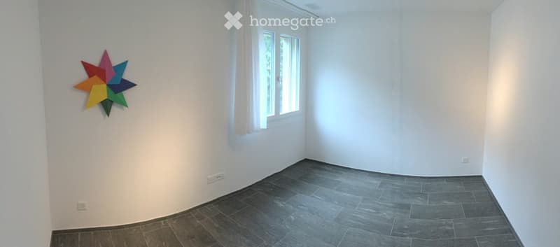 Empty room with white walls, dark grey tiled floor, and a colorful star-shaped wall decoration. Natural light from a window with white curtains.