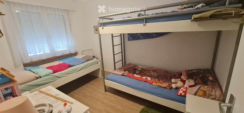 A cozy bedroom with a single bed, a bunk bed, a small table, and a chair. The room features a window with white curtains and a picture frame on the wall.