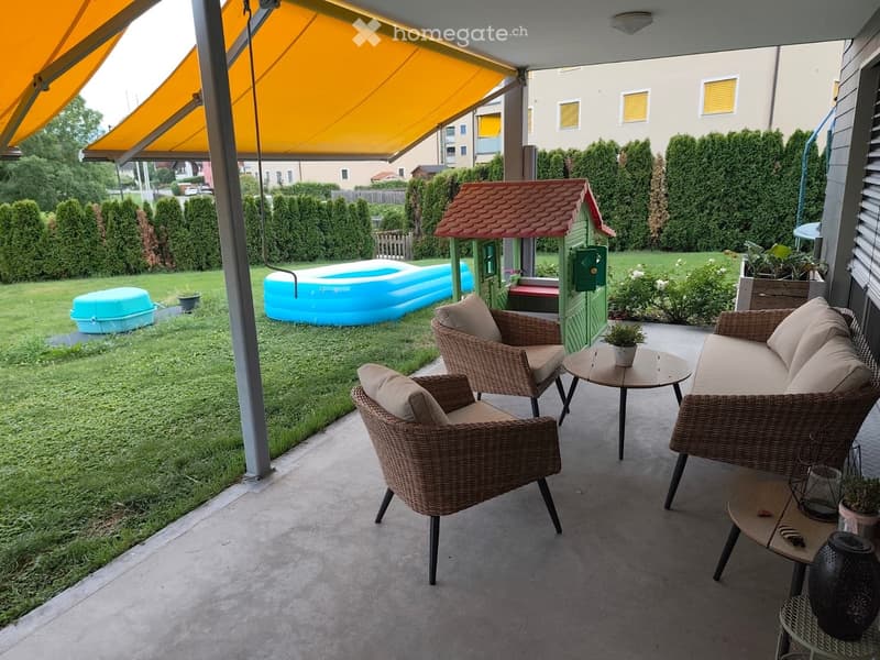 Balcony with wicker furniture, including a sofa, chairs, and tables, with a covered awning and a view of a backyard with a playhouse and inflatable pool.
