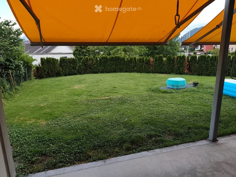 Well-maintained lawn with neatly trimmed hedges, a small blue container, and a covered patio area with orange awnings.