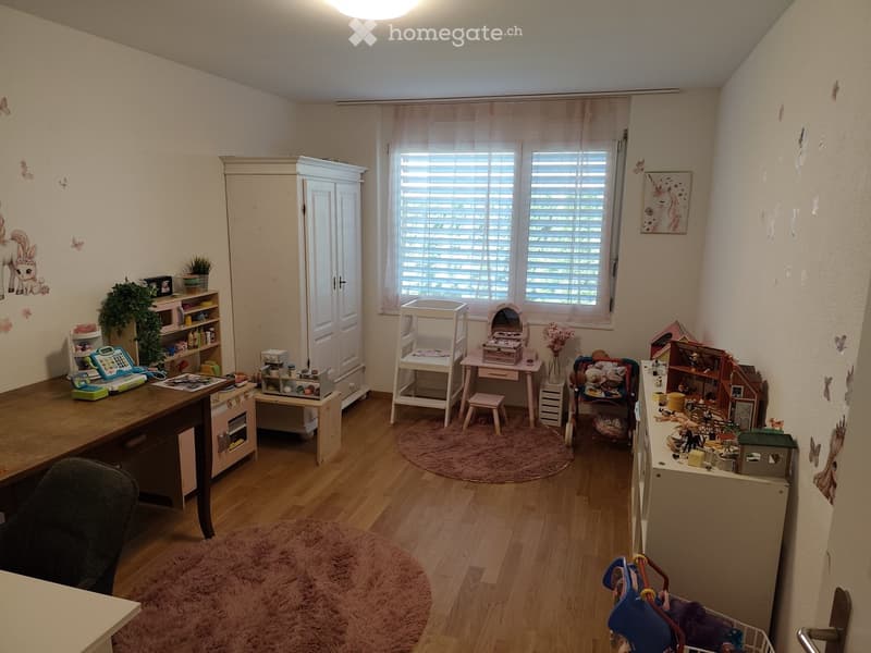 A well-lit room with wooden flooring, a large window with blinds, and various pieces of furniture including a table, chairs, a cabinet, and a small stool. There are toys, a plant, and a mirror on a small table near the window.