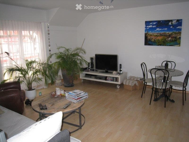 A modern living room with wooden flooring, white walls, and large windows. Features include a sofa, coffee table, dining table with chairs, television, and a potted plant.