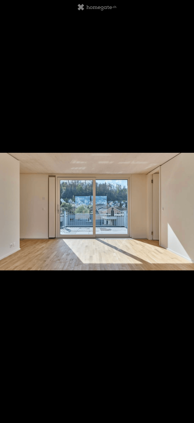 Empty room with wooden flooring, white walls, and large sliding glass doors leading to a balcony with a view of a residential area.