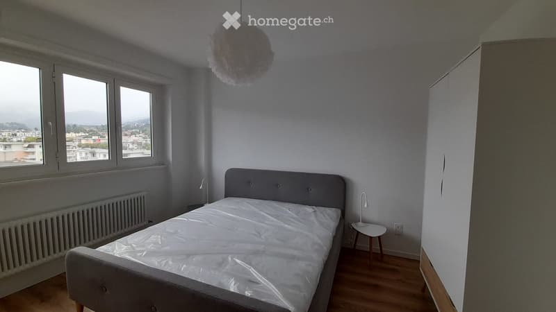 Minimalist bedroom with a grey upholstered bed, white bedding, a small nightstand with a lamp, and large windows offering a city view.