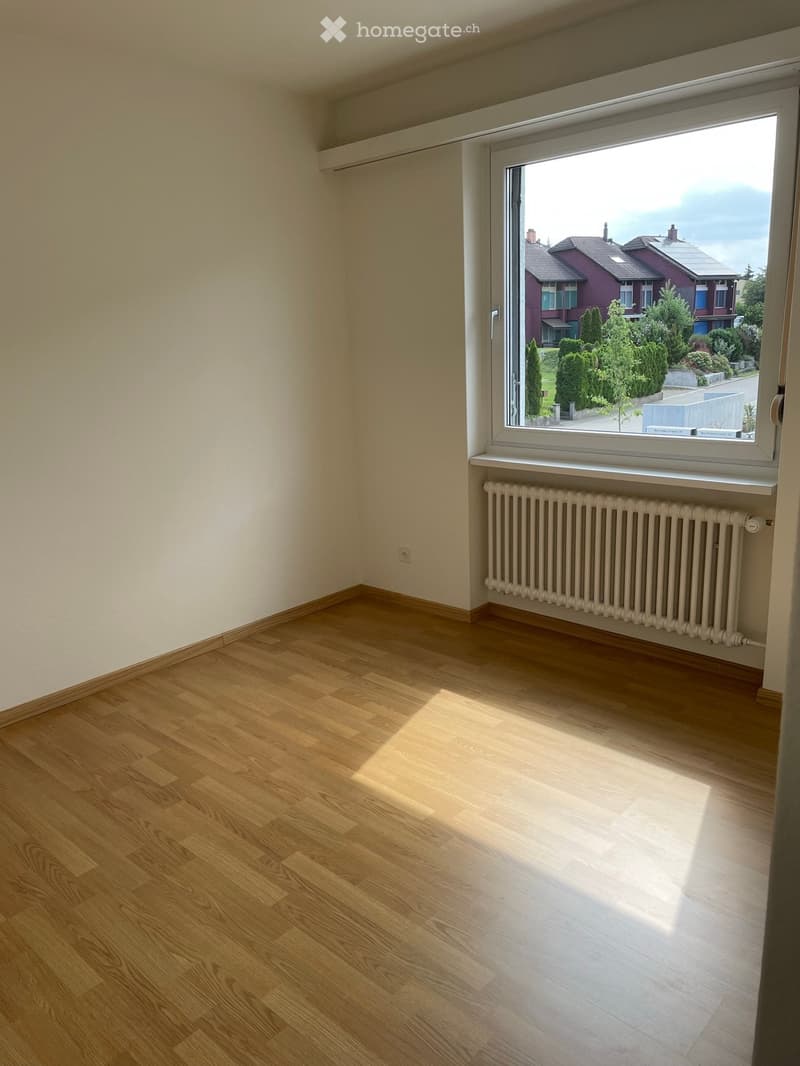 Empty room with wooden flooring, white walls, and a large window showing a view of a residential area with houses and trees.