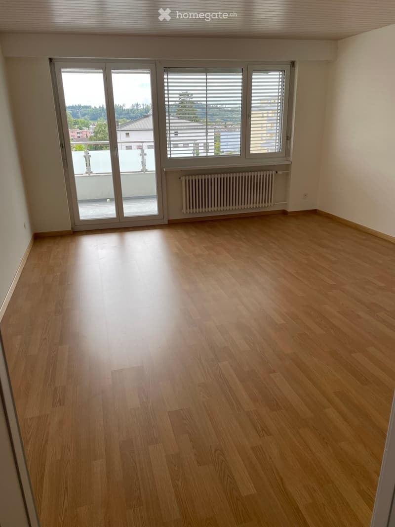 Empty room with wooden flooring, large sliding glass door leading to a balcony, radiator, and window with blinds.