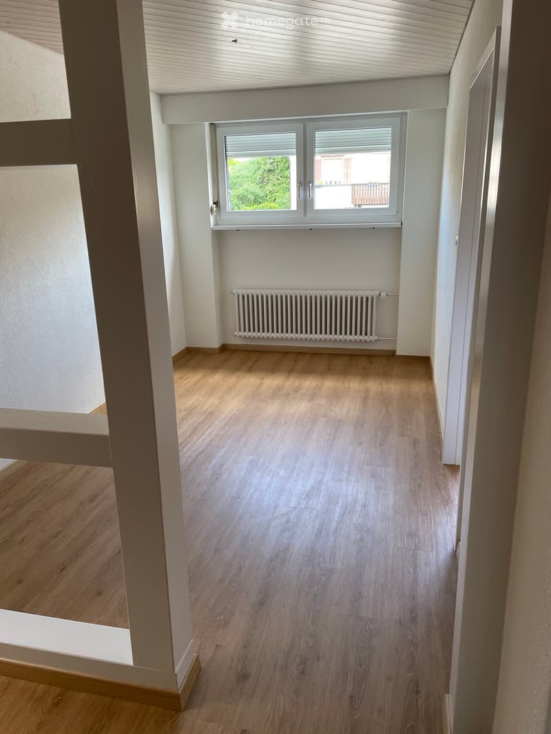 Empty room with wooden flooring, white walls, and a radiator. A window with blinds offers a view of greenery and a balcony.