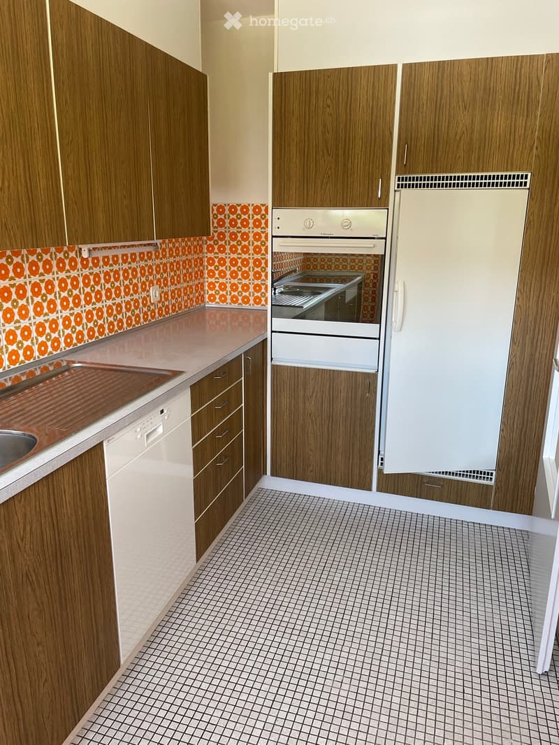 Modern kitchen with wooden cabinets, white appliances, and a tiled backsplash featuring orange circular patterns. The countertop is light-colored, and the floor has a grid-patterned tile design.