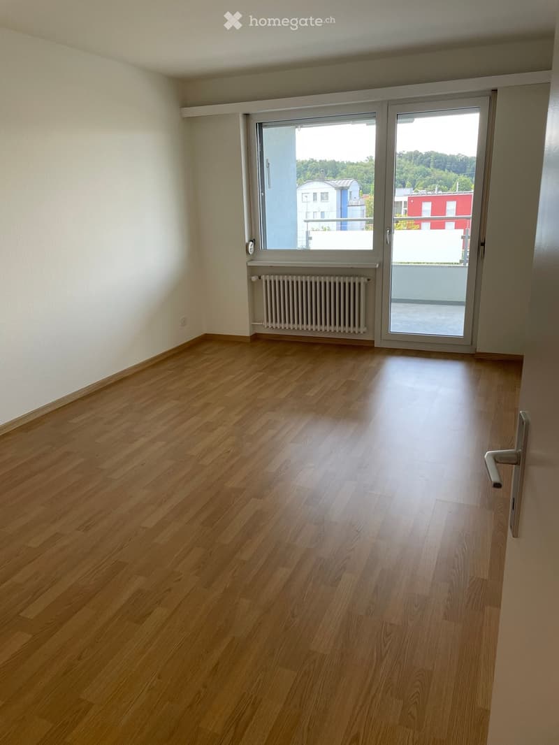 Empty room with wooden flooring, white walls, large window, sliding glass door leading to a balcony, radiator, and visible door handle.