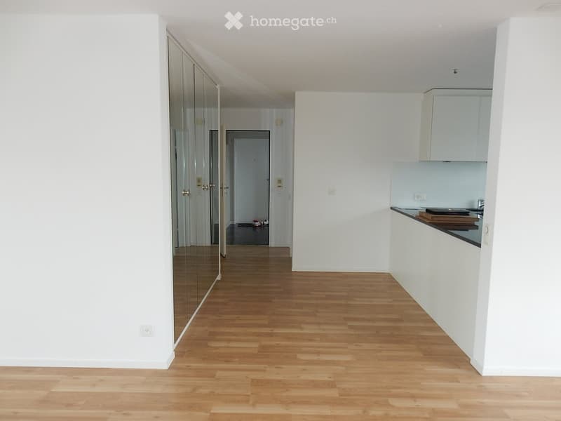 Empty hallway with wooden flooring, white walls, and a small kitchen area visible on the right side.