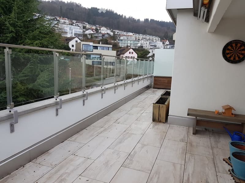Modern balcony with glass railing, tiled floor, wooden bench, small planter, and a view of a town with houses and trees.