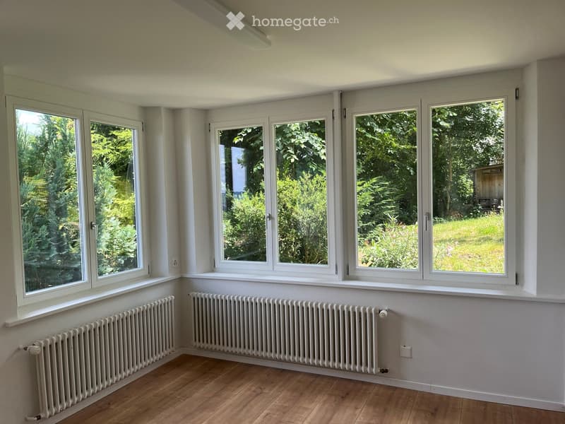 Empty room with wooden flooring, white walls, large windows overlooking a green garden, and two radiators along the wall.