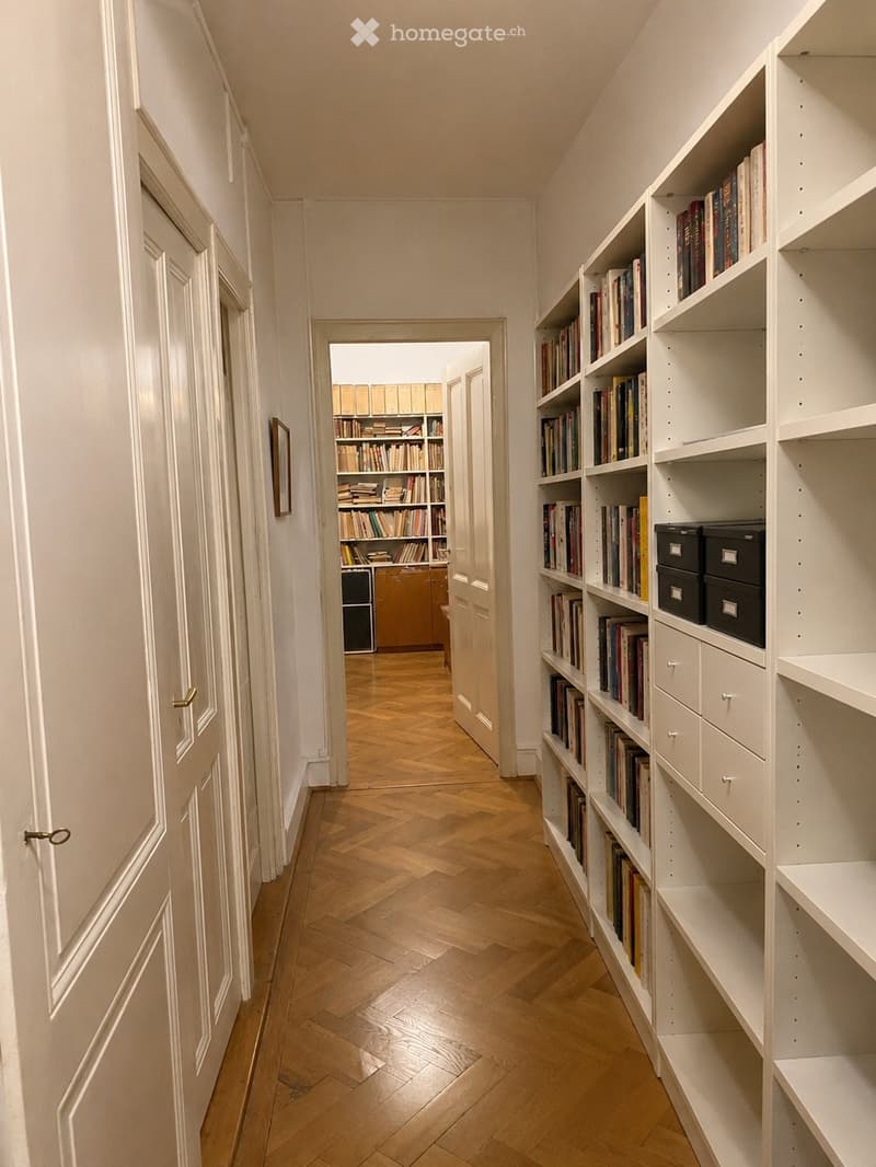 A narrow hallway with wooden flooring and white walls, featuring built-in bookshelves filled with books and storage boxes on the right side. A door at the end leads to another room with more bookshelves.