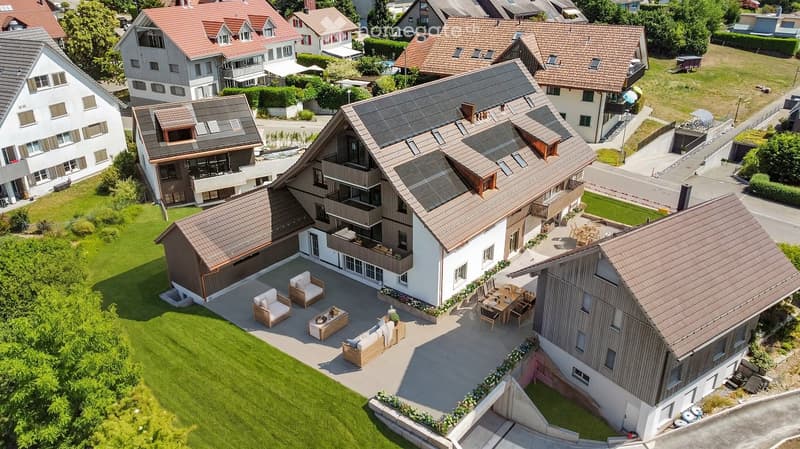 Aerial view of a modern residential complex with multiple houses, featuring a large central house with a spacious patio, outdoor seating, and surrounding greenery.