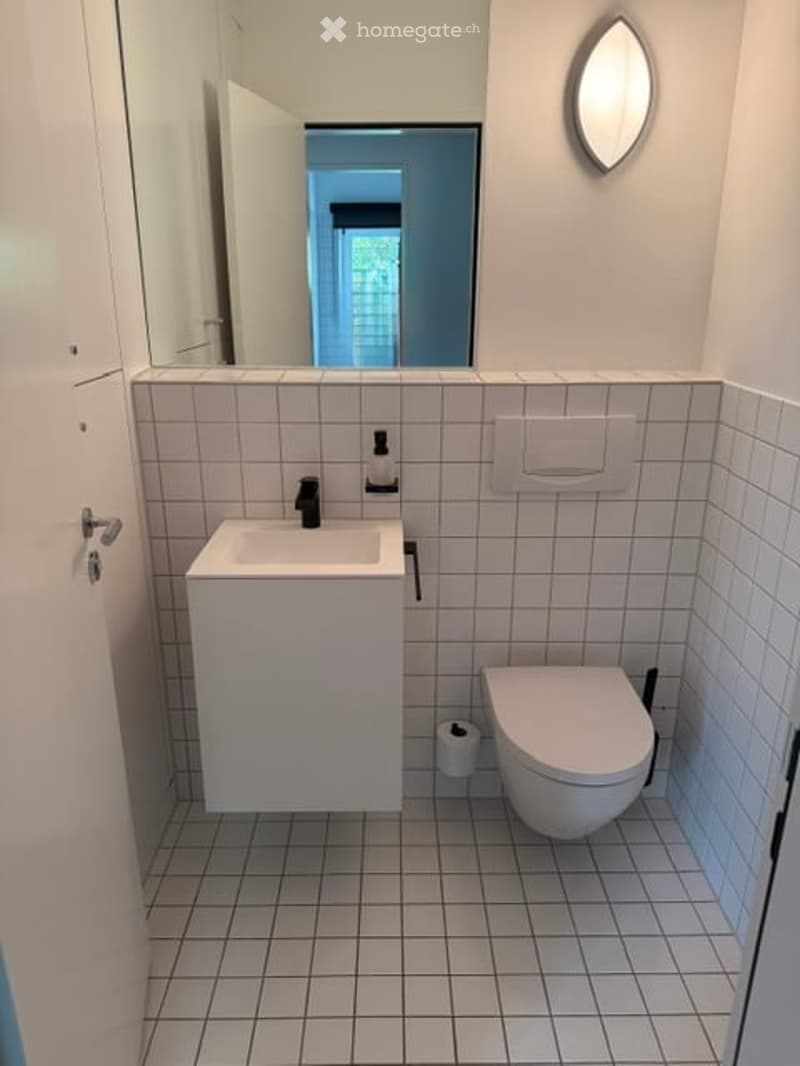 Modern bathroom with white tiled walls and floor, a white sink with a black faucet, a toilet, a round mirror, and a toilet paper holder.