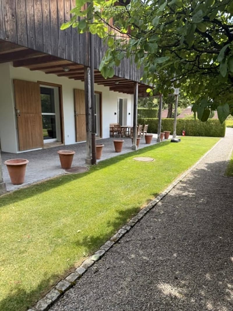 Well-maintained garden area with a gravel pathway, green lawn, and several terracotta pots placed along the pathway. The building in the background features wooden doors and a covered walkway.