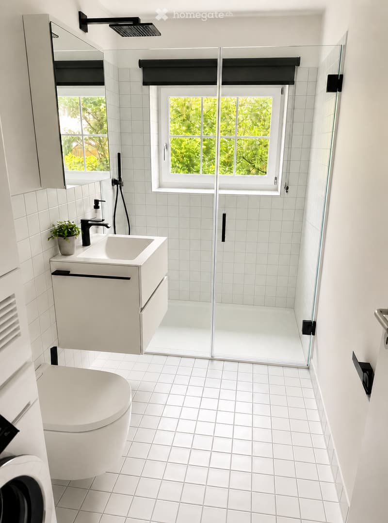 Modern bathroom with white tiled walls and floor, featuring a sink with a faucet, a toilet, a glass-enclosed shower, and a large mirror above the sink.