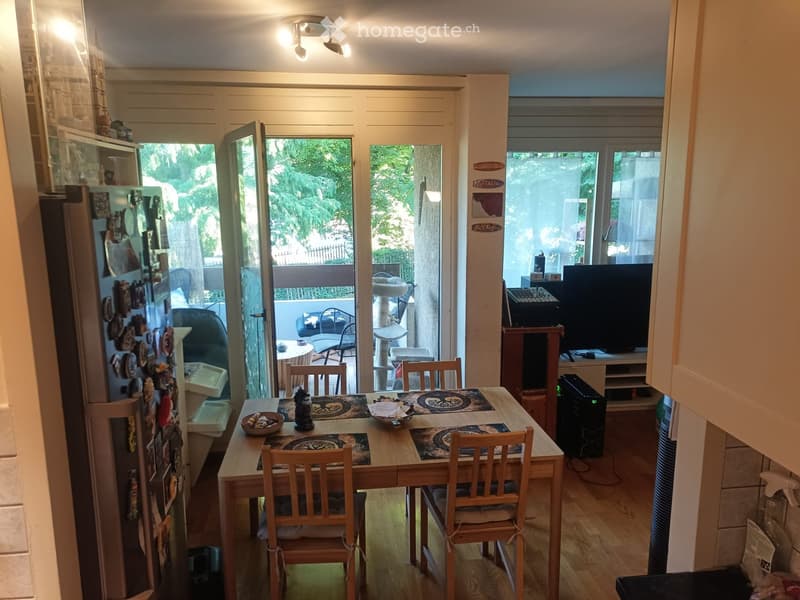 Bright and airy dining area with a wooden dining table, four chairs, a refrigerator, and a television. Large windows and sliding doors open to a balcony with outdoor furniture.