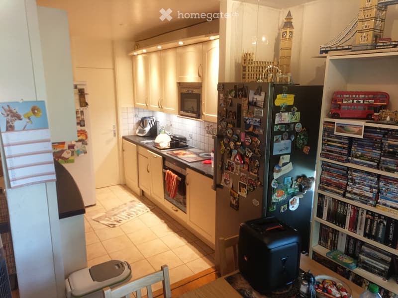 A modern kitchen with white cabinets, stainless steel appliances, and a refrigerator covered with magnets and notes. A dining area with a table, chairs, and a shelf filled with books and DVDs is visible.