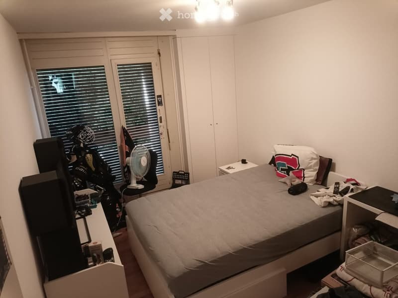 A small, furnished bedroom with a bed, a fan, a chair, a laptop, a screen, a table, and a storage unit. The room has white walls, a large window with blinds, and a closet.