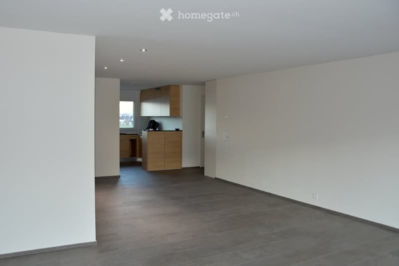 Empty room with wooden flooring, white walls, and a small kitchen area visible in the background.