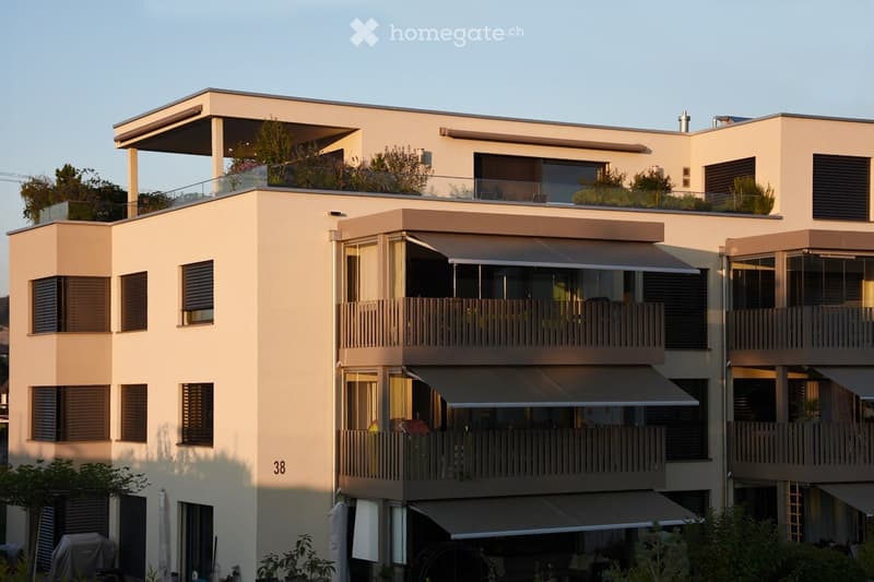 Modern multi-story residential building with balconies, large windows, and a rooftop terrace with greenery.