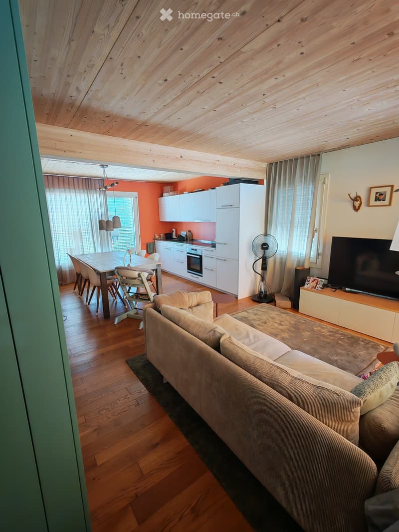 Open-plan living and dining area with wooden floors, a beige sofa, a dining table with chairs, a white kitchen with cabinets and an oven, and a fan near the window.