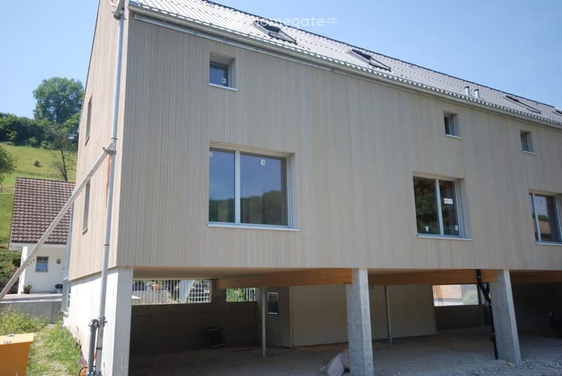 Modern two-story house with raised ground floor, light wood siding, large windows, and a covered garage area.