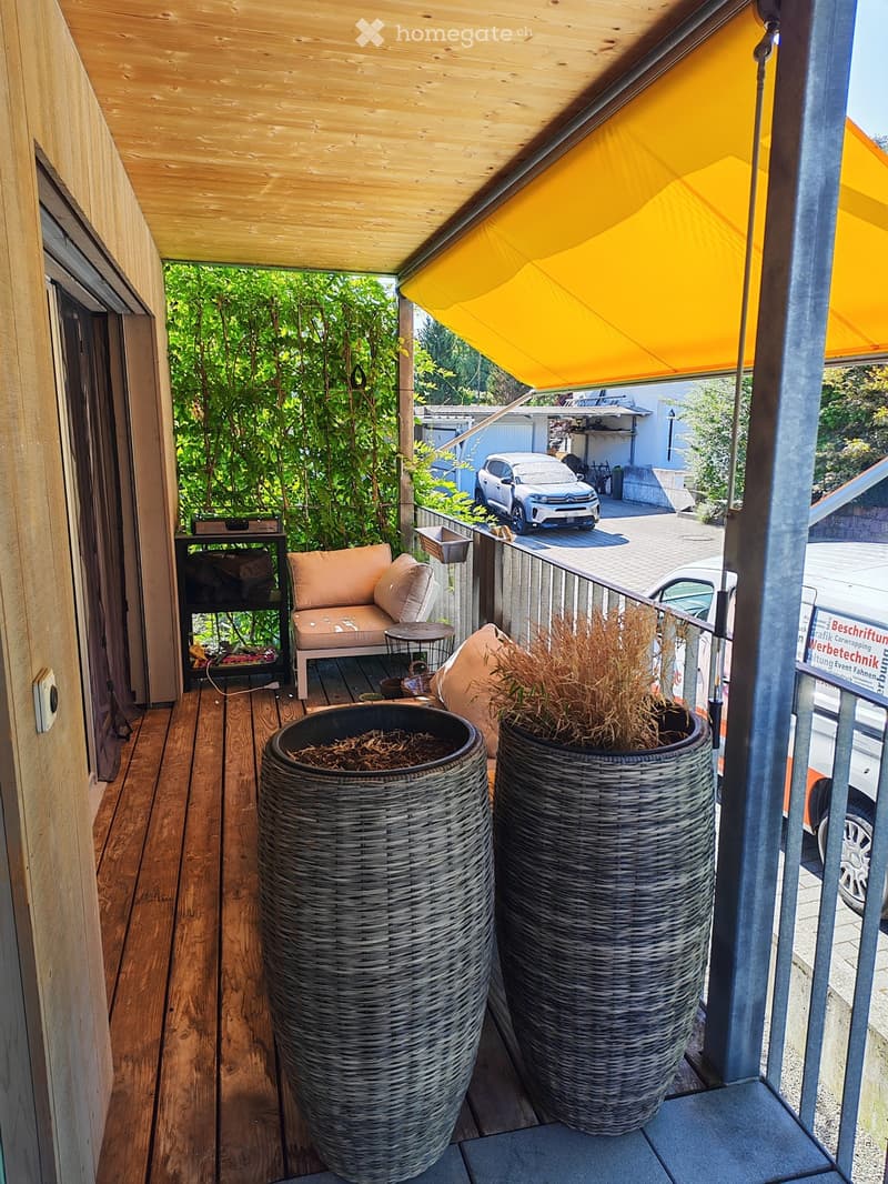 Balcony with wooden decking, covered by a yellow awning, featuring a sofa, small table, and two large wicker planters.