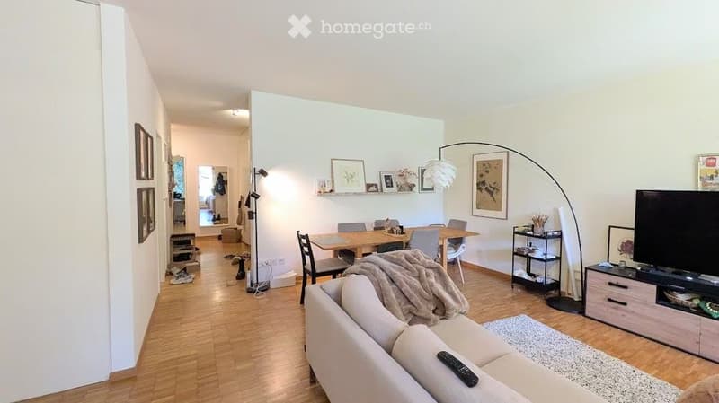 Bright living and dining area with wooden flooring, a beige sofa, a coffee table, a dining table with chairs, a floor lamp, shelves, and a television.
