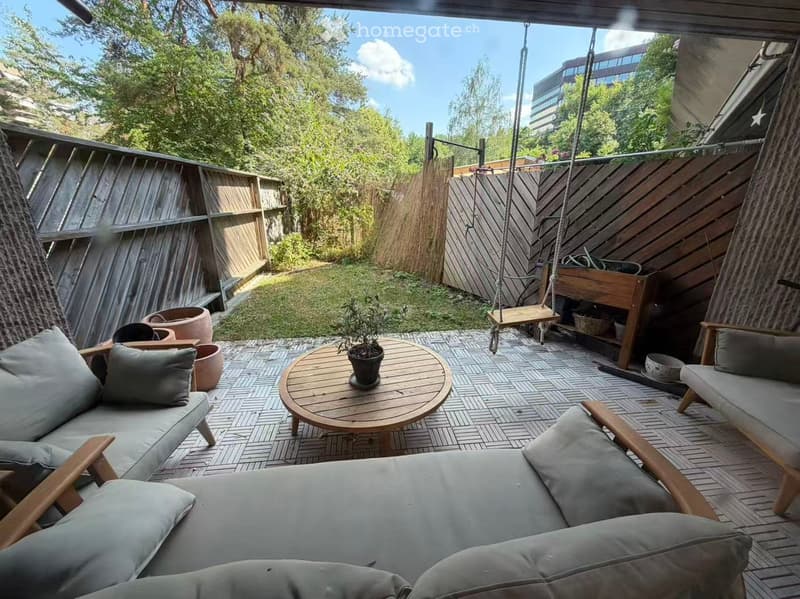 A wooden decked balcony with a round wooden table, potted plant, two sofas with cushions, and a hanging swing. The area is enclosed with wooden fencing and features outdoor seating.