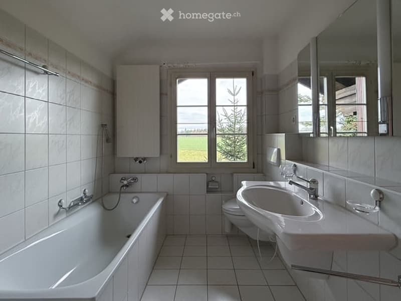 Bright bathroom with white tiles, a bathtub, a sink, a toilet, and a large window offering a view of greenery.
