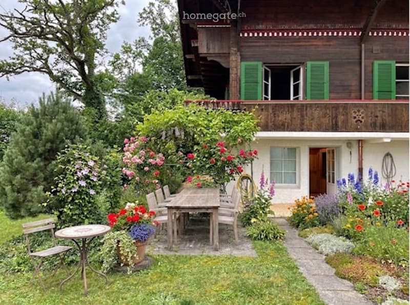 A well-maintained garden area with a wooden table and chairs, surrounded by colorful flowers and greenery, leading to a wooden house with green shutters and a balcony.