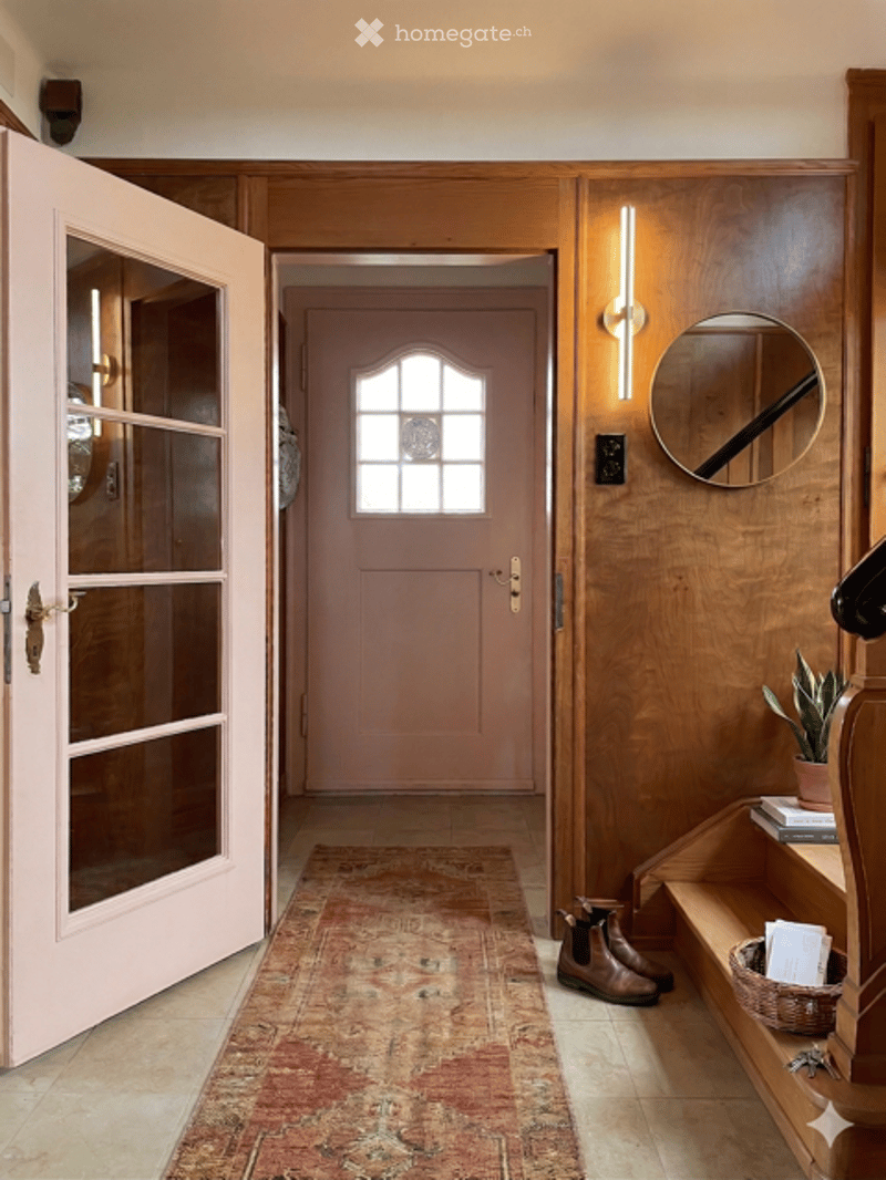 Elegant hallway with wooden paneling, a pink front door, a patterned rug, and a small shelf with a plant and books.