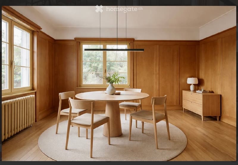 Dining room with wooden panel walls, large windows, round dining table with six chairs, sideboard with lamp, and a vase with greenery on the table.
