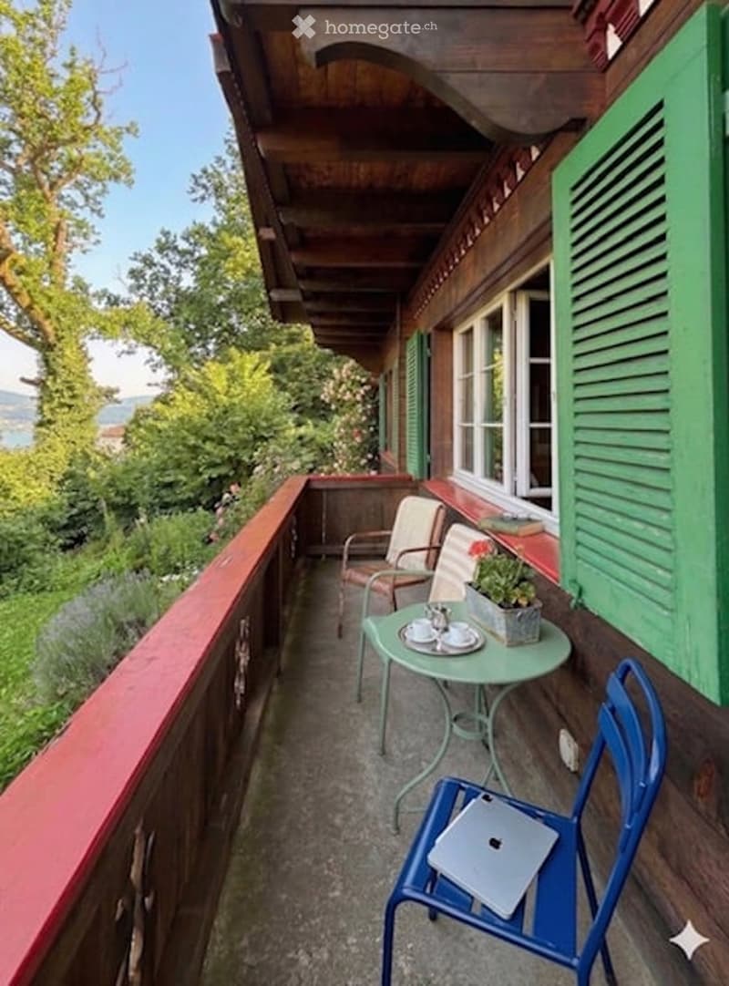 Balcony with wooden beams, green shutters, and a scenic view of trees and mountains. Includes a small round table with chairs and a laptop.