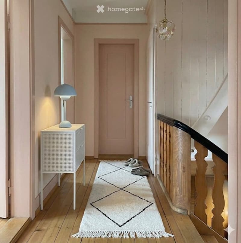 A narrow hallway with light pink walls, wooden flooring, a white woven cabinet with a blue lamp, a mirror, a closed door, a white runner rug with geometric patterns, and a staircase with wooden railings.