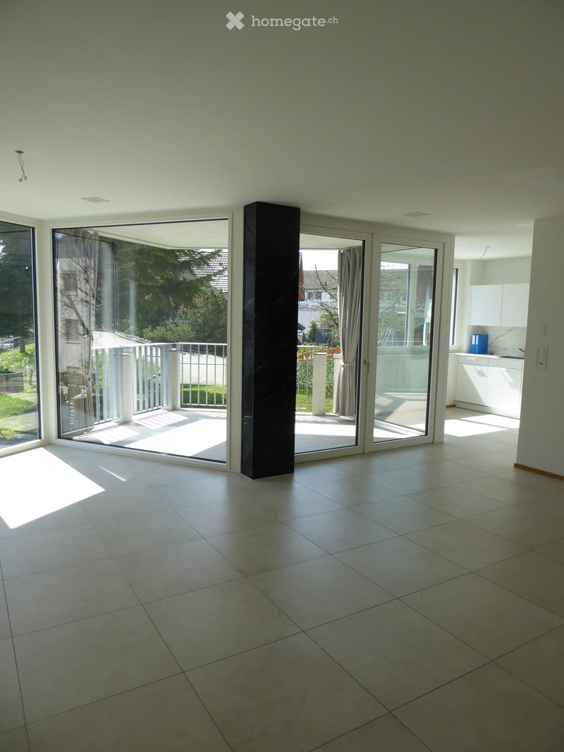 Bright, empty room with large sliding glass doors leading to a balcony, tiled floor, and modern white cabinetry visible in the adjacent area.
