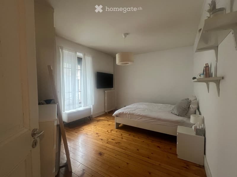 Cozy bedroom with wooden flooring, a bed with white bedding, a nightstand with a lamp, a television mounted on the wall, and a window with white curtains.