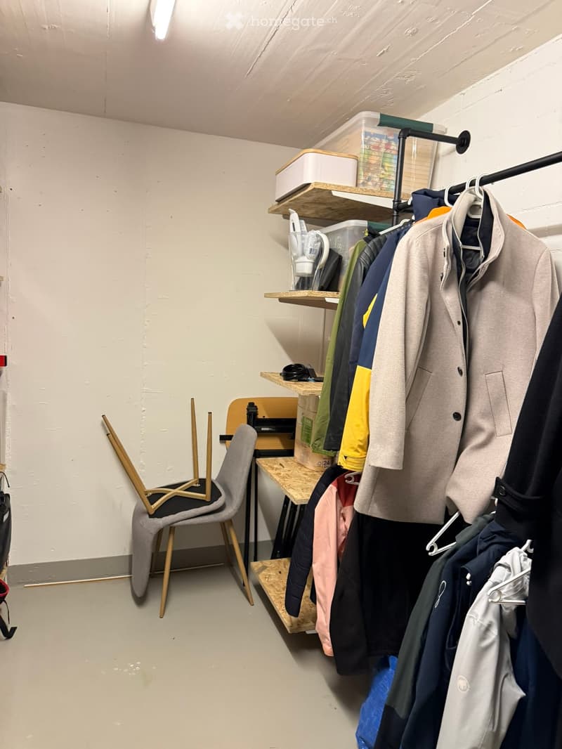 A small, furnished room with a grey chair, a wooden table, shelves holding various items, and a clothing rack with multiple coats and jackets.