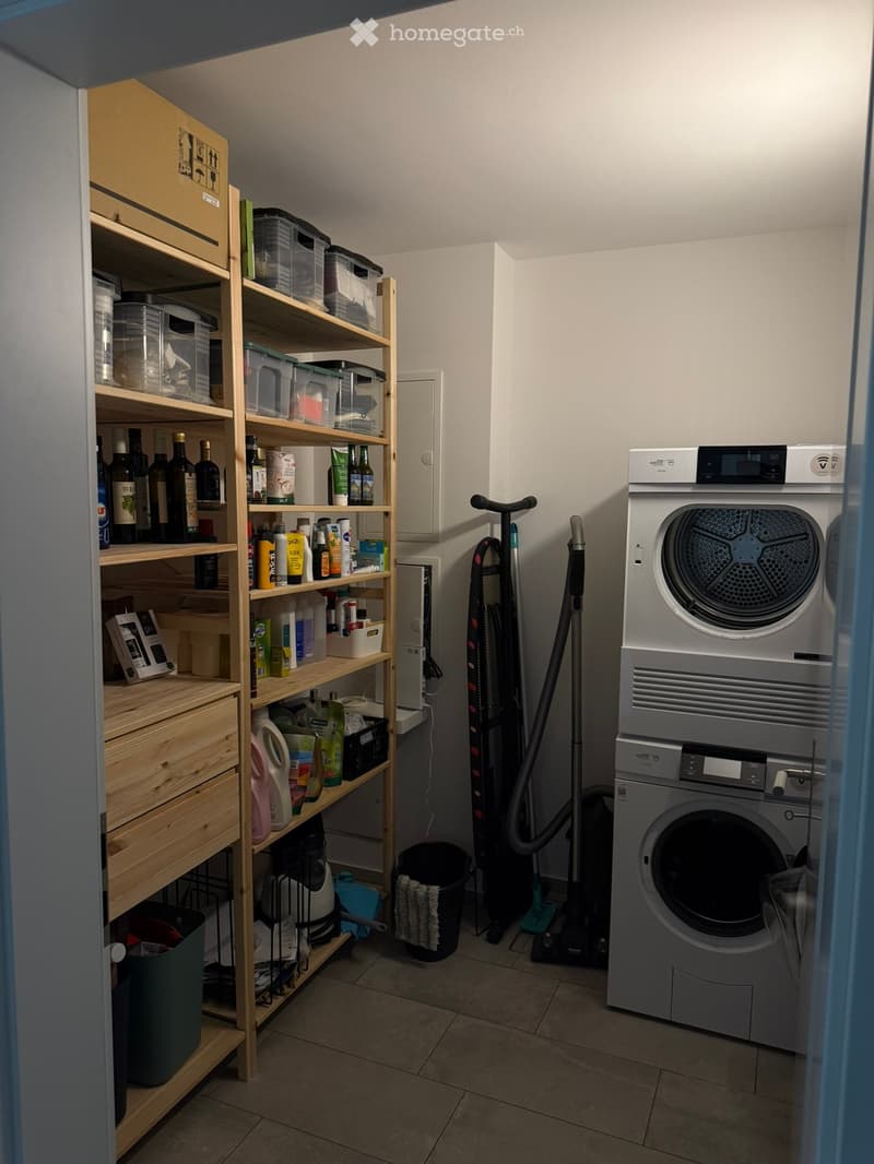 A small utility room with a washing machine, drying machine, wooden shelf filled with bottles and cleaning supplies, vacuum cleaner, and a bin.
