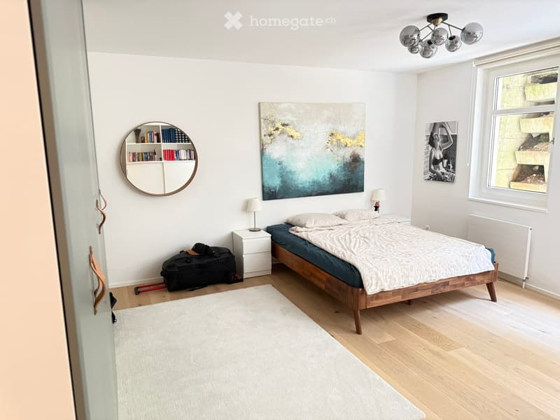 Cozy bedroom with a wooden bed, white nightstand, round mirror, bookshelf, and artwork on the walls. The room features light wooden flooring and a white rug.