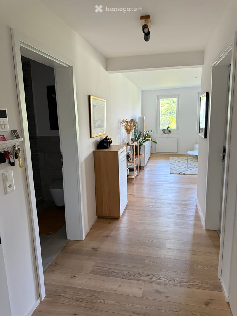 Bright hallway with wooden flooring, white walls, and several doors leading to different rooms. Features include a small wooden console table with a vase, a framed picture on the wall, and a coat rack with keys and bags.