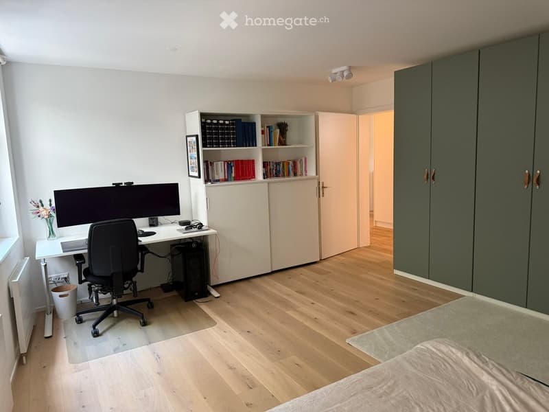 Modern, well-lit room with a desk setup, a bed, and built-in storage. Features include a large screen, ergonomic chair, bookshelf, and a green wardrobe.