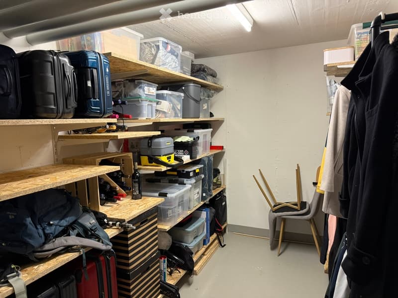 Storage room with wooden shelves holding suitcases, backpacks, wine bottles, and various storage boxes. A chair and coats are visible on the right side.
