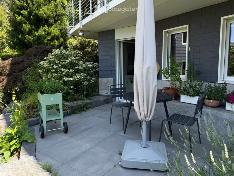 Outdoor patio area with a table, chairs, umbrella, and a small cart, surrounded by plants and flowers, with a modern house in the background.