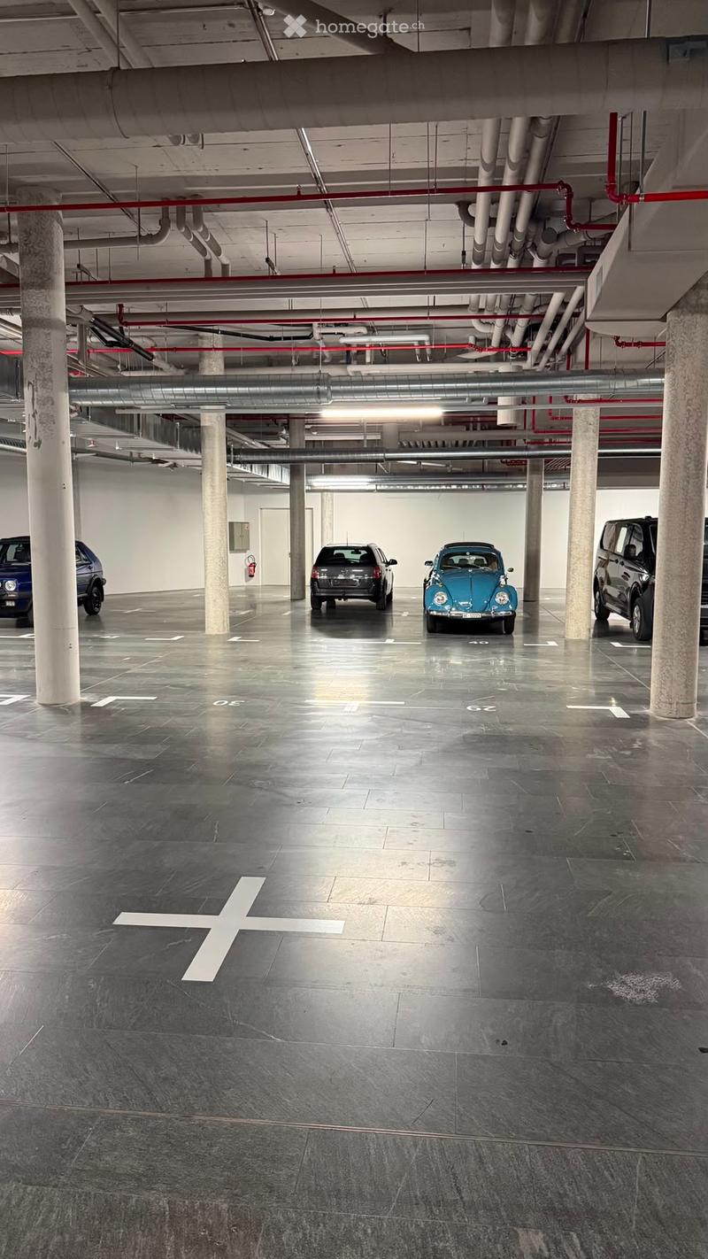 Modern underground parking area with several parked cars, concrete pillars, and exposed pipes.