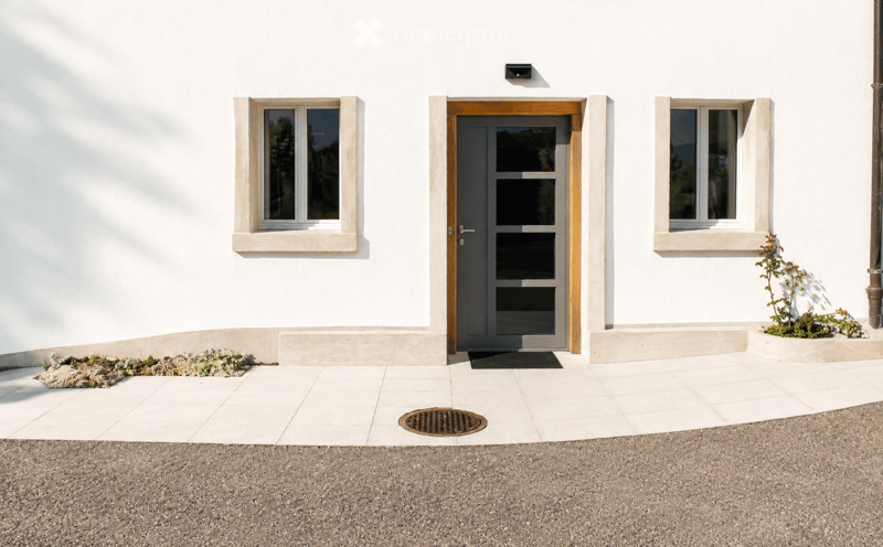 White house exterior with a central grey door framed by wooden trim, flanked by two windows with white frames and stone sills. The entrance has a raised concrete platform and a small garden bed with plants to the left.