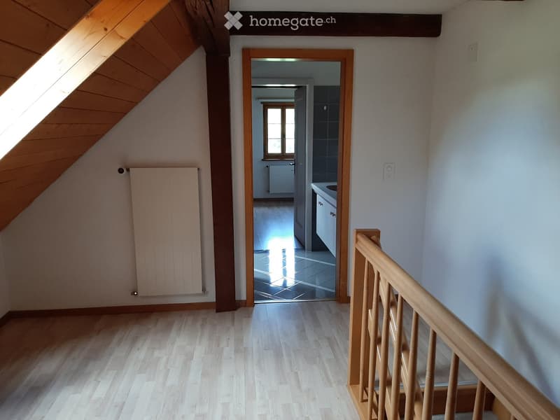 Bright hallway with wooden flooring, white walls, and a wooden railing leading to a room with a window and a radiator.
